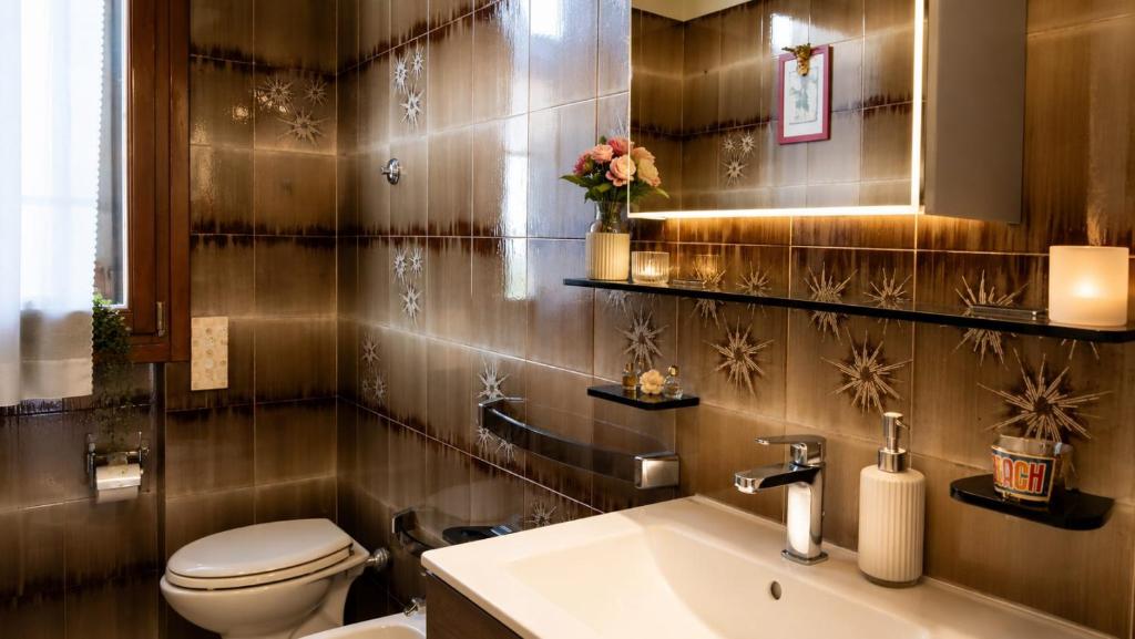 A bathroom with a white sink and toilet, dark patterned tiled walls, shelves, and warm lighting.